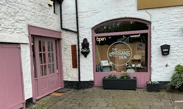 White building with pink doors and a sign for 'The Artisan's Den' on a street.